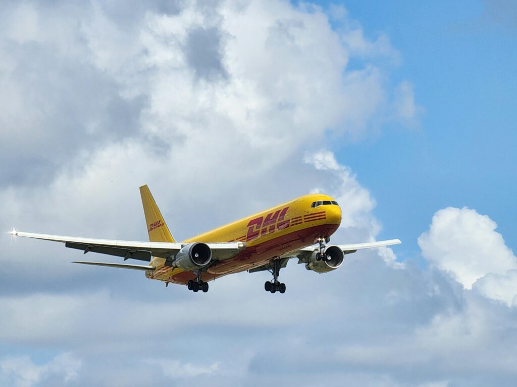 A DHL cargo plane in flight under a bright blue sky filled with fluffy clouds.